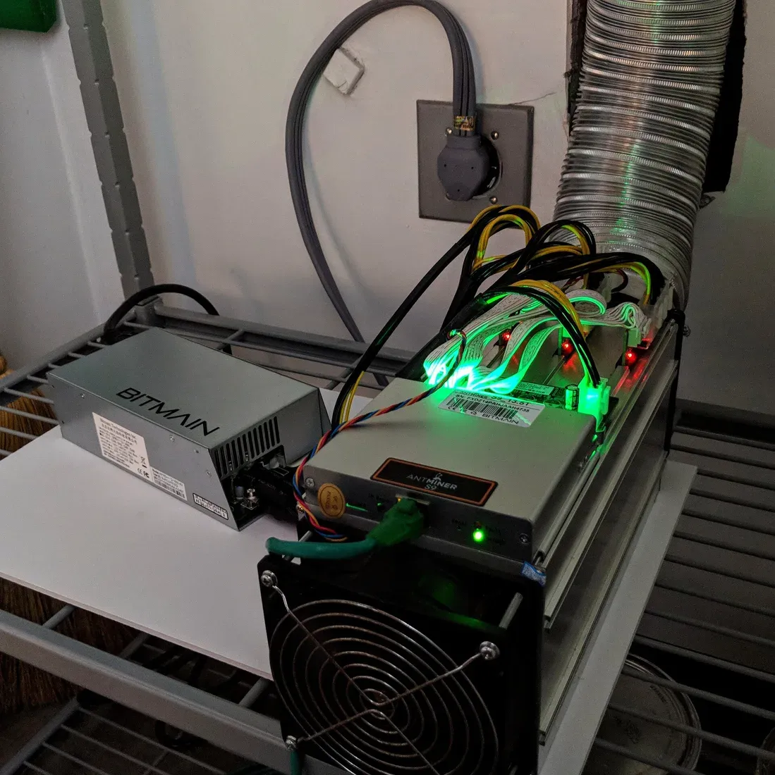 جهاز تعدين Antminer S9 فضي اللون بمروحة كبيرة في الأمام، وأسلاك طاقة كثيرة تخرج من أعلاه إلى مزود طاقة منفصل مكتوب عليه BITMAIN، وأنبوب تهوية معدني مرن متصل بالجهاز من الخلف
