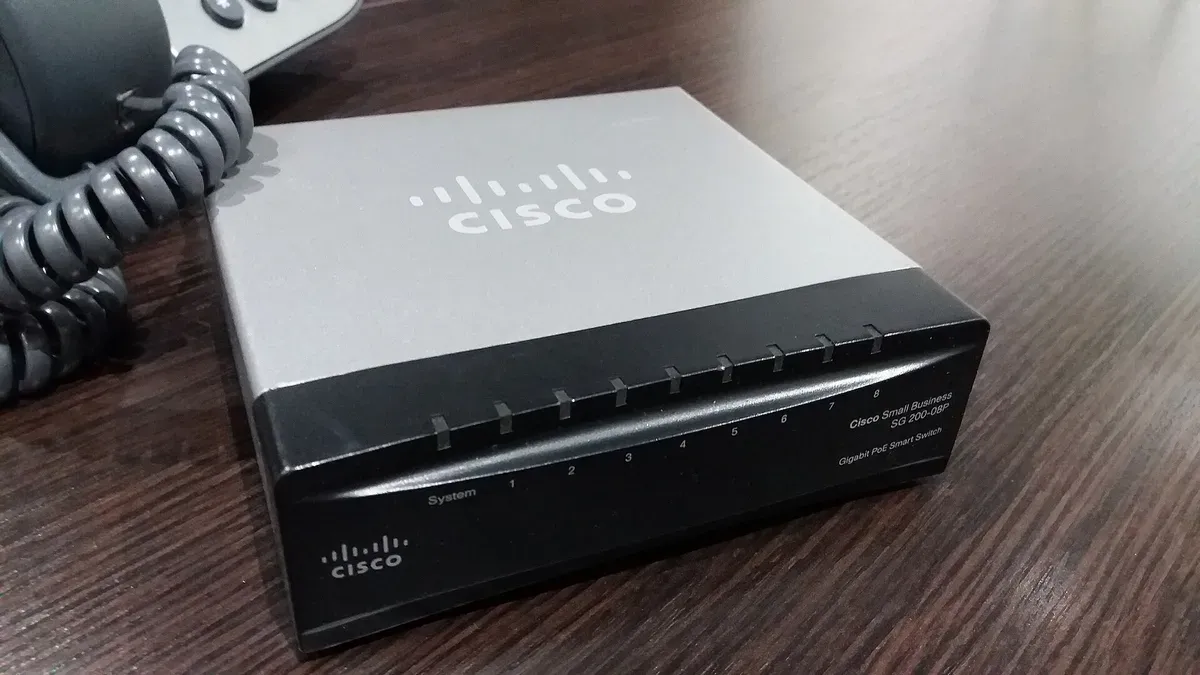 Switch صغير من Cisco بغطاء رمادي ومقدمة سوداء على مكتب خشبي، مكتوب عليه Cisco Small Business SG 200-08P Gigabit PoE Smart Switch، ومصابيح لثمانية منافذ مرقمة ومصباح System، وبجانبه سلك هاتف ملفوف