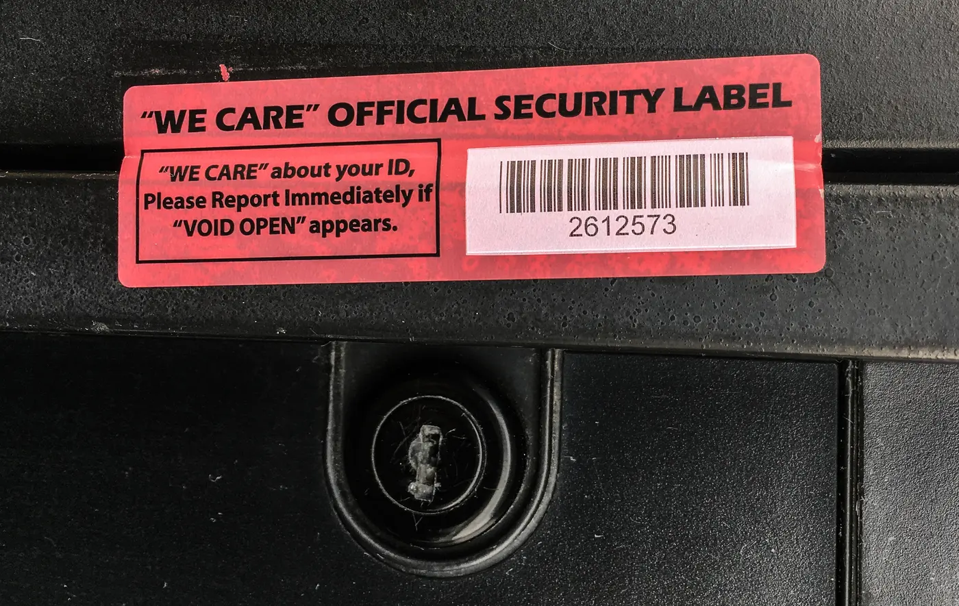 ملصق أحمر على باب جهاز دفع في مضخة وقود مكتوب عليه Official Security Label وعليه رقم تسلسلي وباركود وطلب الإبلاغ إذا ظهرت عبارة VOID OPEN، وتحته قفل الباب