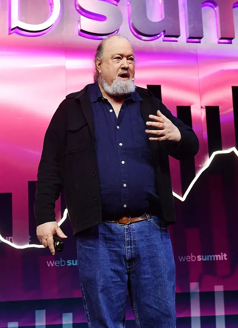 David Chaum على مسرح مؤتمر يرتدي سترة سوداء وقميصاً أزرق ويتحدث بيده اليمنى، وخلفه شاشة وردية عليها شعار المؤتمر