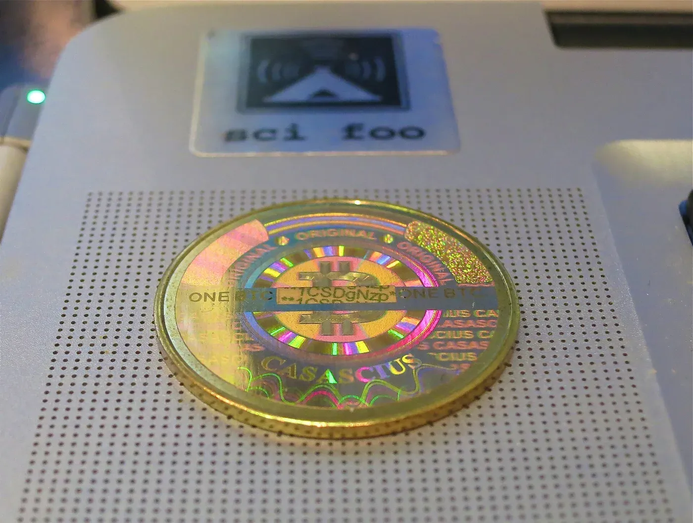 عملة معدنية ذهبية اللون عليها ملصق ثلاثي الأبعاد ملون عليه شعار Bitcoin وكلمة CASASCIUS وجزء من عنوان مطبوع، موضوعة على سطح جهاز رمادي