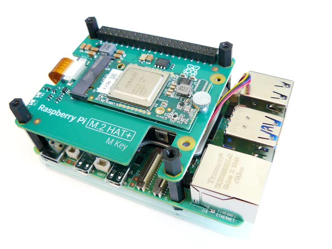 لوحة Raspberry Pi 5 خضراء وفوقها لوحة M.2 HAT+ عليها وحدة Hailo بشريحة فضية مكتوب عليها HAILO، مع منافذ USB والشبكة على جانب اللوحة