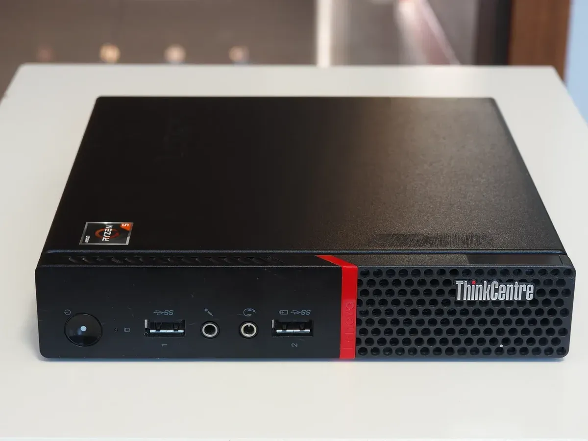 حاسوب Lenovo ThinkCentre M715q أسود صغير بحجم الكتاب على طاولة بيضاء، في واجهته زر التشغيل ومنفذا USB ومنفذا الصوت وفتحات تهوية مع شعار ThinkCentre وخط أحمر