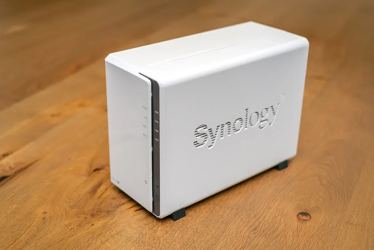 جهاز Synology DiskStation أبيض بقرصين على طاولة خشبية، على جانبه شعار Synology محفور، وفي واجهته مؤشرات STATUS وLAN وDISK 1 وDISK 2 وزر التشغيل