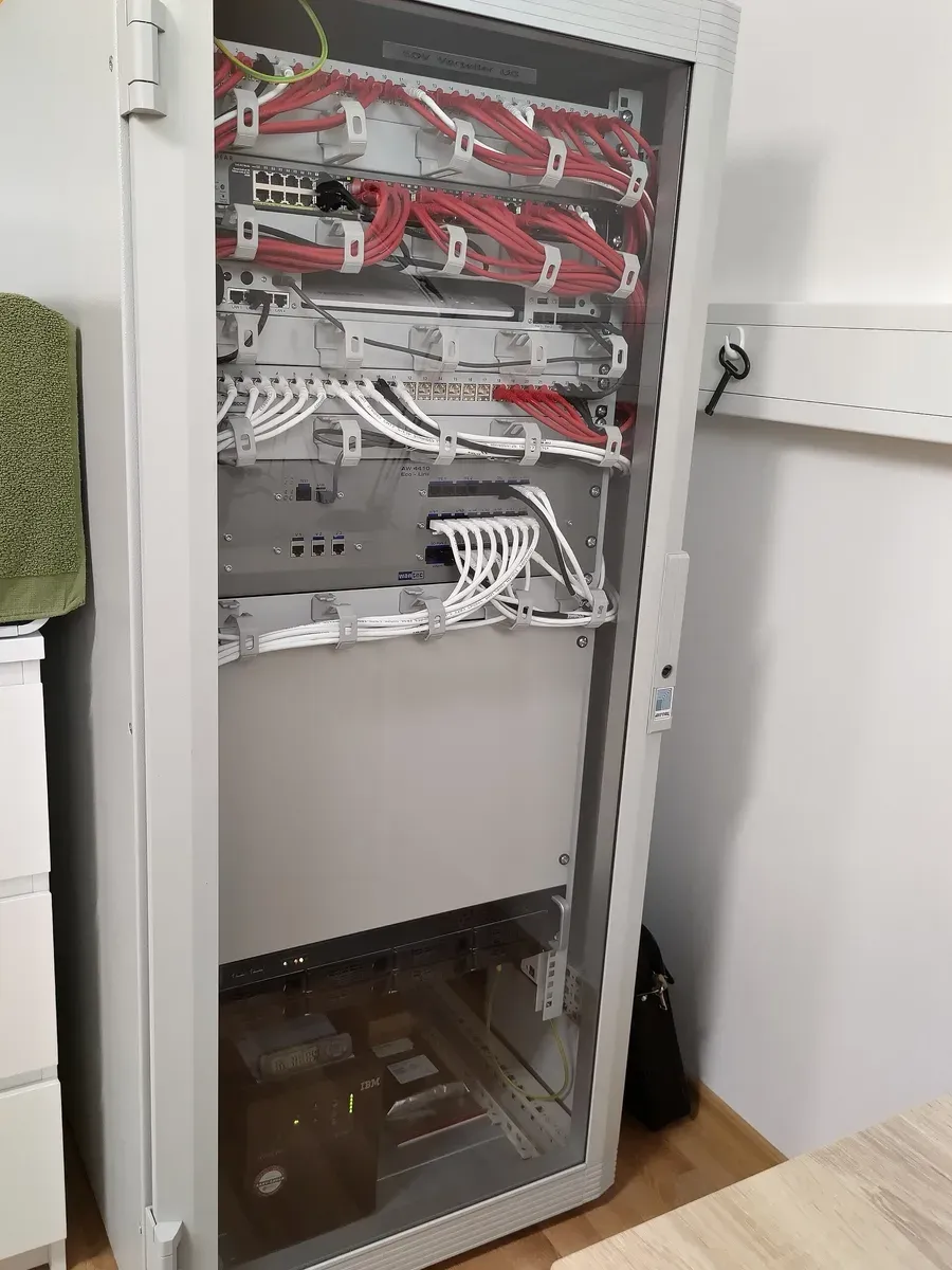 خزانة شبكة رمادية بباب زجاجي في غرفة بيت، فيها من الأعلى Patch panels وSwitch بكابلات حمراء وبيضاء مرتبة في حلقات لتنظيم الكابلات، ثم أجهزة بمنافذ زرقاء، ثم لوح فارغ، وفي الأسفل سيرفر