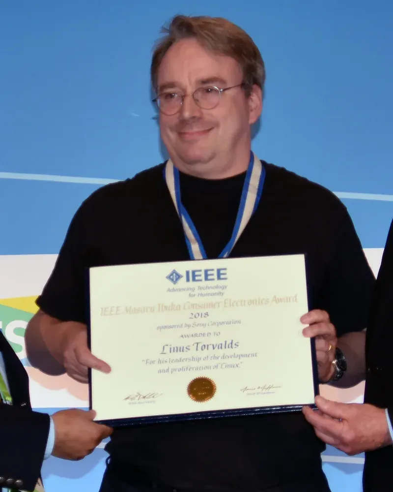 لينوس تورفالدز يبتسم ويحمل شهادة جائزة IEEE Masaru Ibuka للإلكترونيات الاستهلاكية لعام 2018 مكتوباً عليها اسمه، ويرتدي قميصاً أسود ووشاح الميدالية حول عنقه