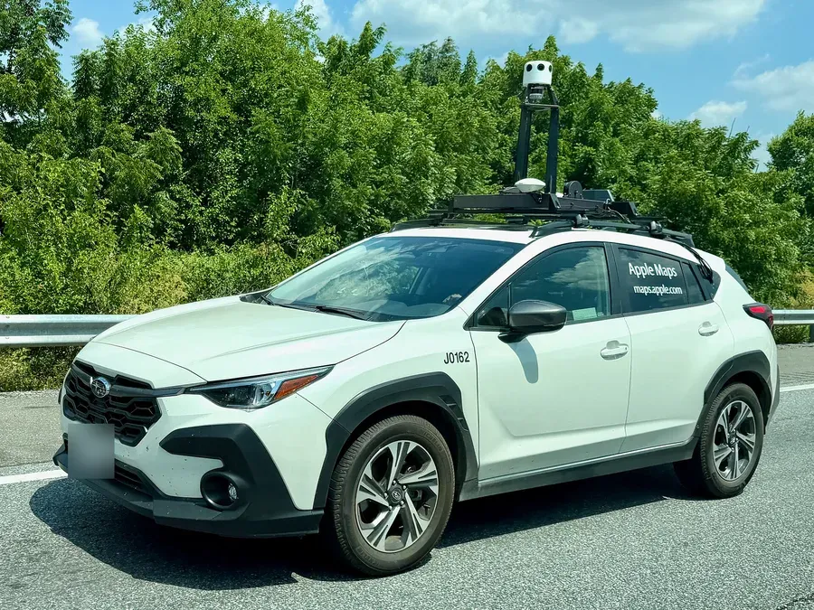 سيارة Subaru بيضاء على طريق سريع مكتوب على زجاجها الخلفي Apple Maps وmaps.apple.com، وفوق سقفها حامل أسود يحمل عموداً في أعلاه وحدة بيضاء أسطوانية