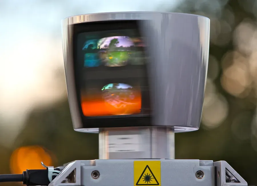 جهاز LiDAR فضي أسطواني من Velodyne يظهر ضبابياً من سرعة دورانه، وفيه نافذتان داكنتان تعكسان الأشجار والسماء، وتحته قاعدة رمادية عليها علامة تحذير ليزر صفراء