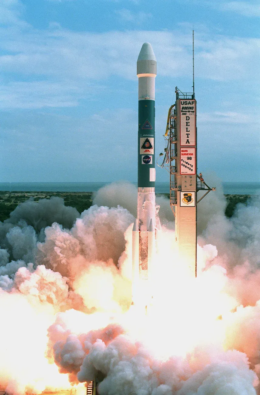 صاروخ Delta II يرتفع عن منصة الإطلاق وسط سحابة كبيرة من الدخان واللهب، وعلى برج الإطلاق لوحة مكتوب عليها Mars Surveyor 98 Climate Orbiter