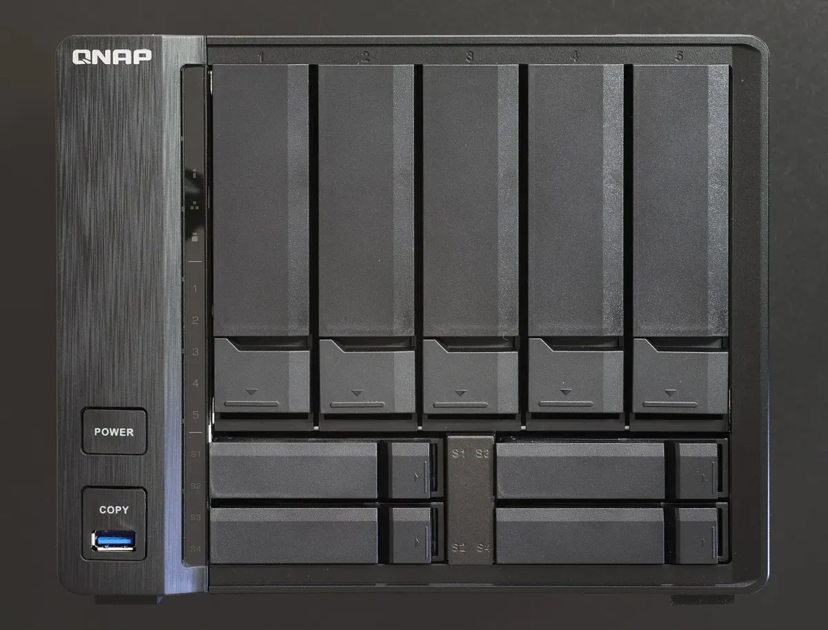 واجهة جهاز QNAP TVS-951X أسود، في يساره شعار QNAP وزر التشغيل وزر النسخ بجانب منفذ USB أزرق، وفي يمينه خمسة أدراج عمودية مرقمة من 1 إلى 5 للأقراص الكبيرة، وتحتها أربعة أدراج أفقية صغيرة مرقمة S1 إلى S4