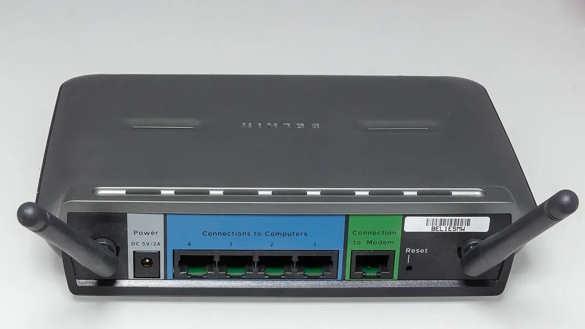ظهر Router منزلي رمادي من Belkin بهوائيين، فيه أربعة منافذ داخل مستطيل أزرق مكتوب عليه Connections to Computers، ومنفذ واحد داخل مستطيل أخضر مكتوب عليه Connection to Modem، ومنفذ الكهرباء وزر Reset