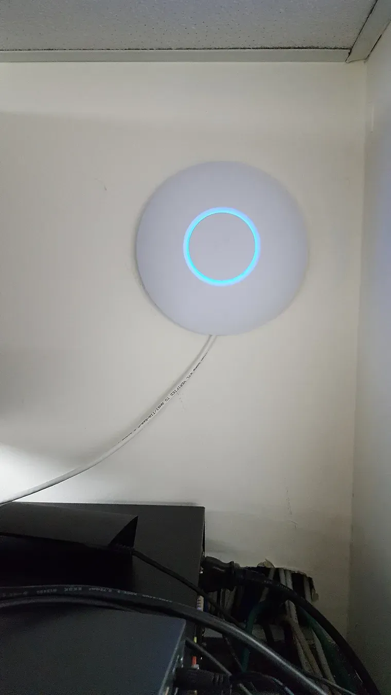 نقطة وصول دائرية بيضاء من UniFi مثبتة على سقف بجوار الجدار، في وسطها حلقة ضوء زرقاء، ويخرج منها كابل Ethernet واحد أبيض