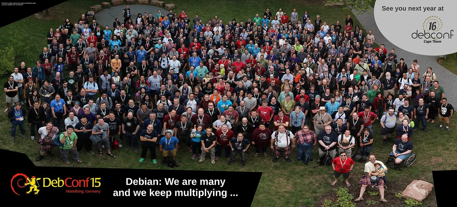صورة جماعية من الأعلى لمئات المشاركين في DebConf15 واقفين على العشب، وعليها شعار DebConf15 وعبارة Debian: We are many and we keep multiplying