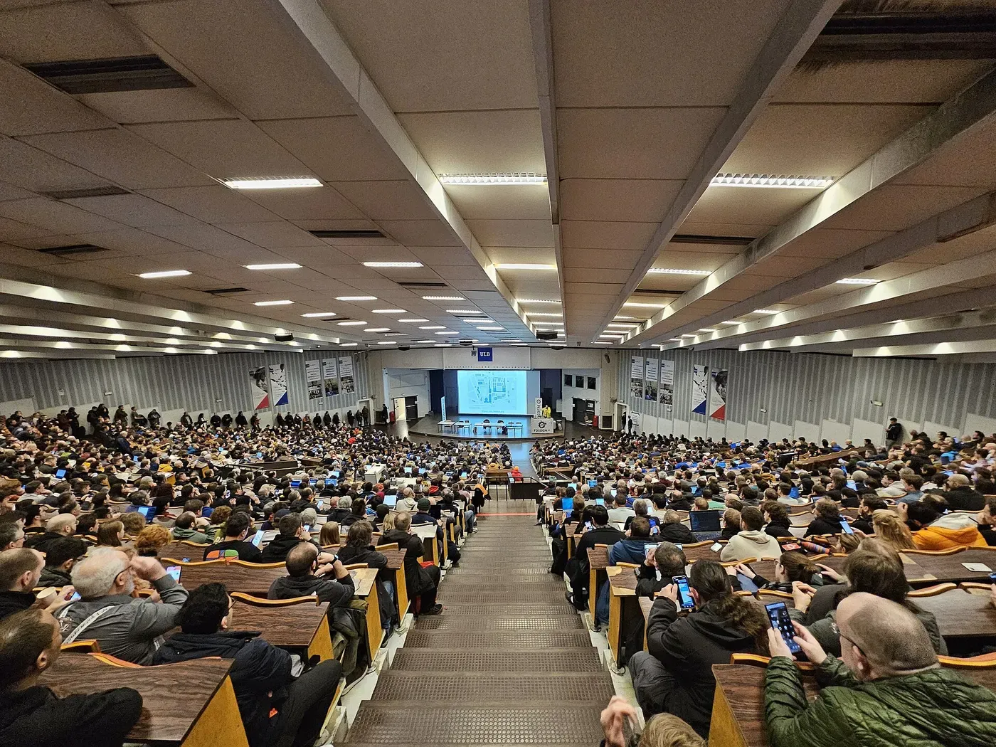 قاعة محاضرات كبيرة متدرجة ممتلئة بمئات الحضور أمام شاشة ومنصة في الأسفل، وعلى الجدار شعار جامعة ULB