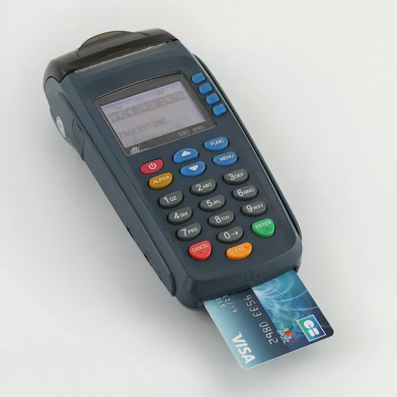 جهاز نقاط بيع تقليدي بشاشة صغيرة ولوحة أرقام بأزرار ملونة، وبطاقة Visa مدخلة في فتحة الشريحة أسفل الجهاز