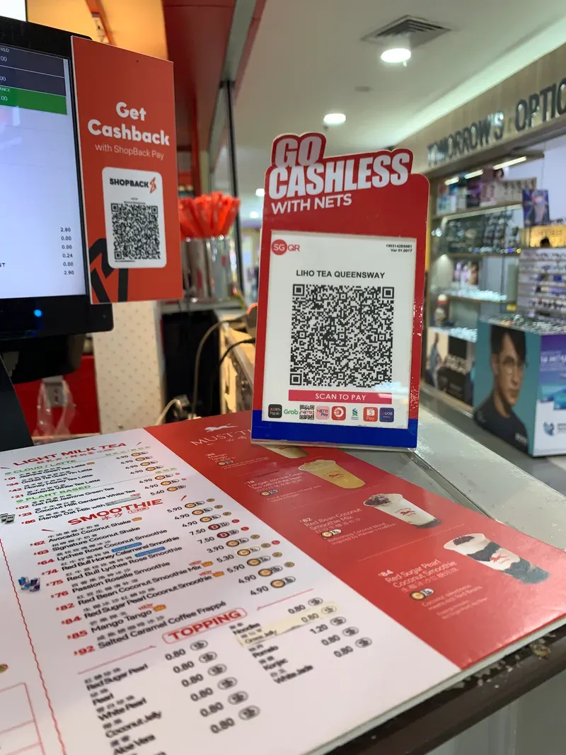 لوحة مطبوعة على طاولة الدفع في محل مشروبات فيها رمز QR كبير وتحته شعارات تطبيقات الدفع التي تقبله، وبجانبها قائمة الأسعار